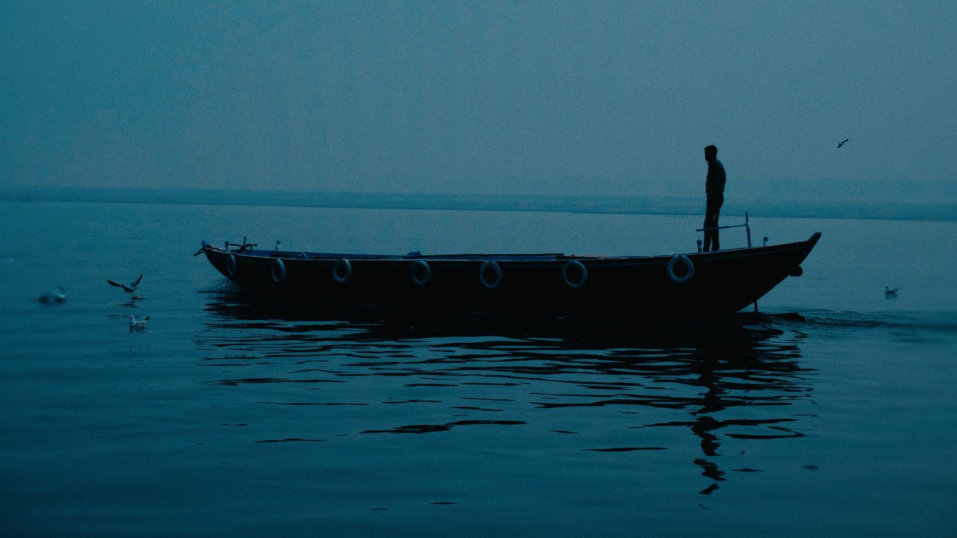 Un barcaiolo in piedi all’alba sul Gange a Varanasi, in controluce sull’acqua immobile
