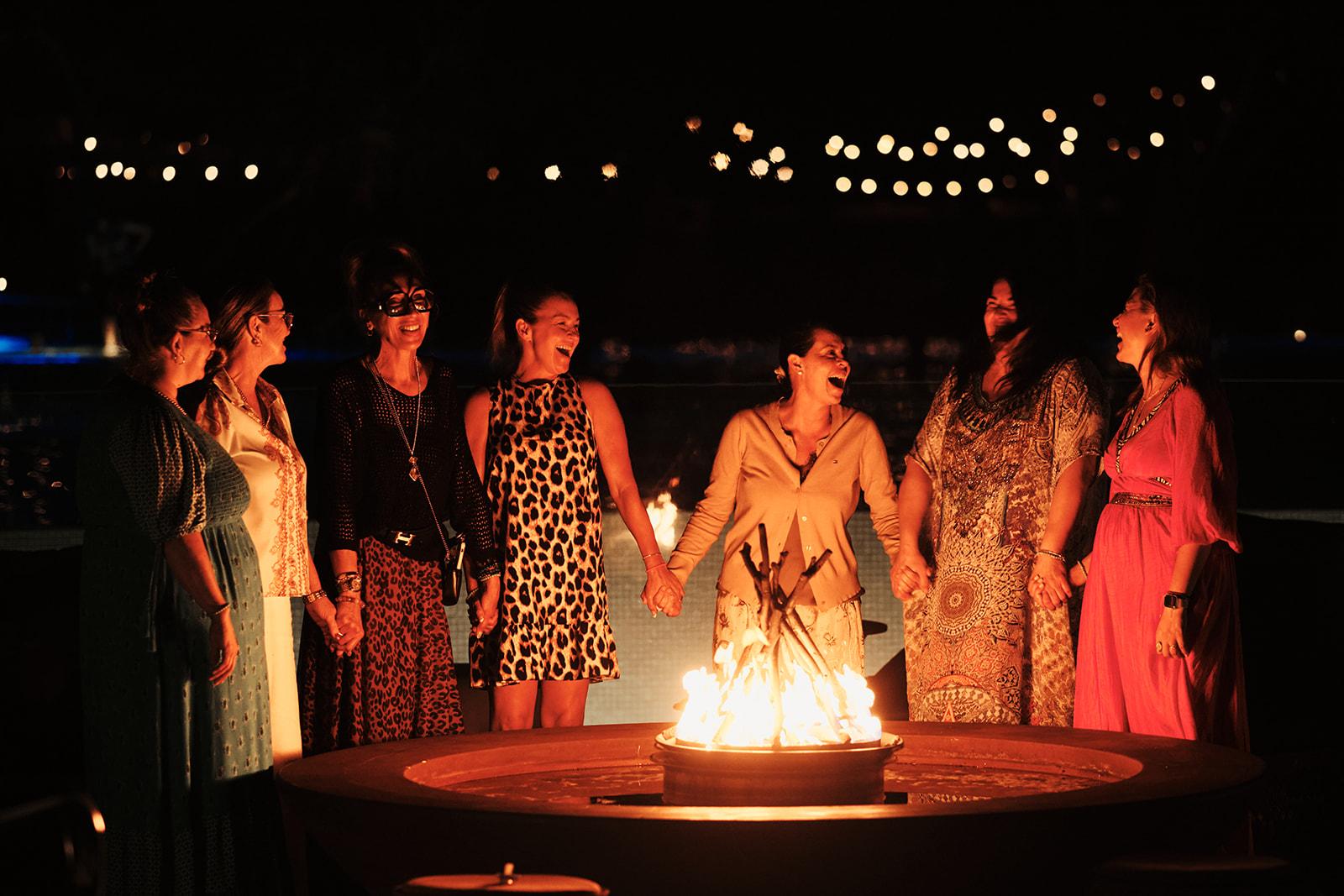 A circle of women holding hands and laughing around a lit fire pit at night