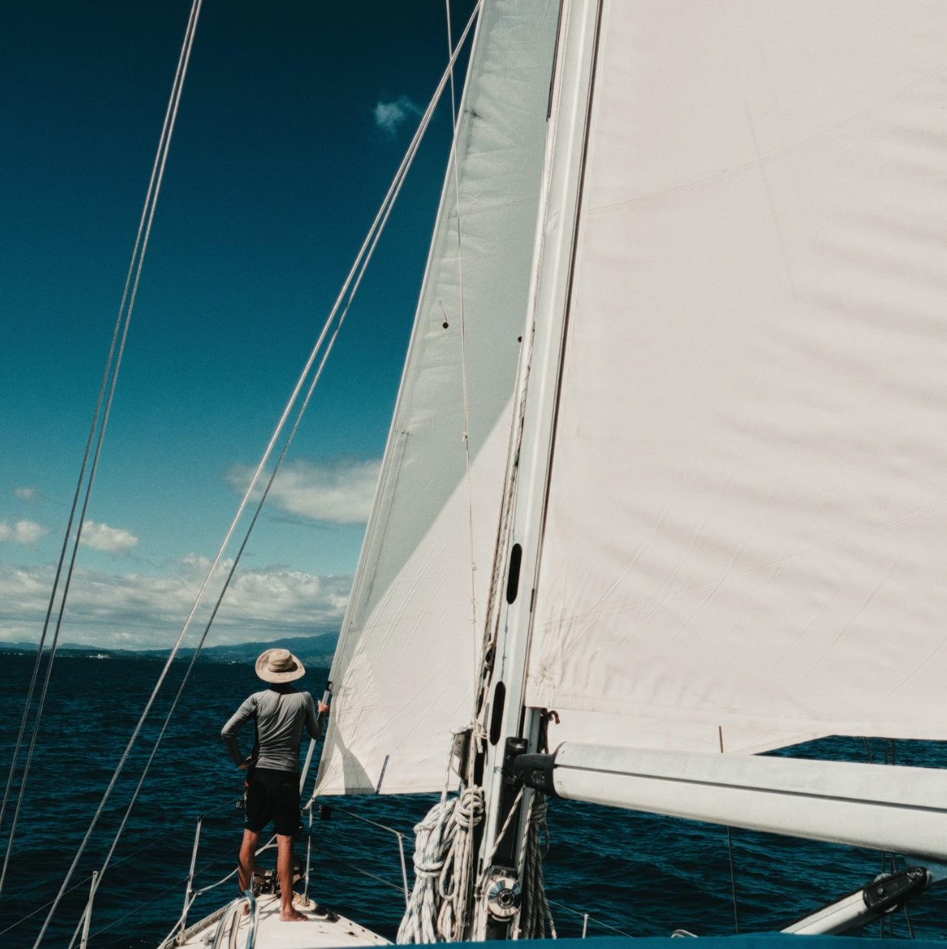 Nharyan standing at the bow, hand on the mast, sails full against a blue sky