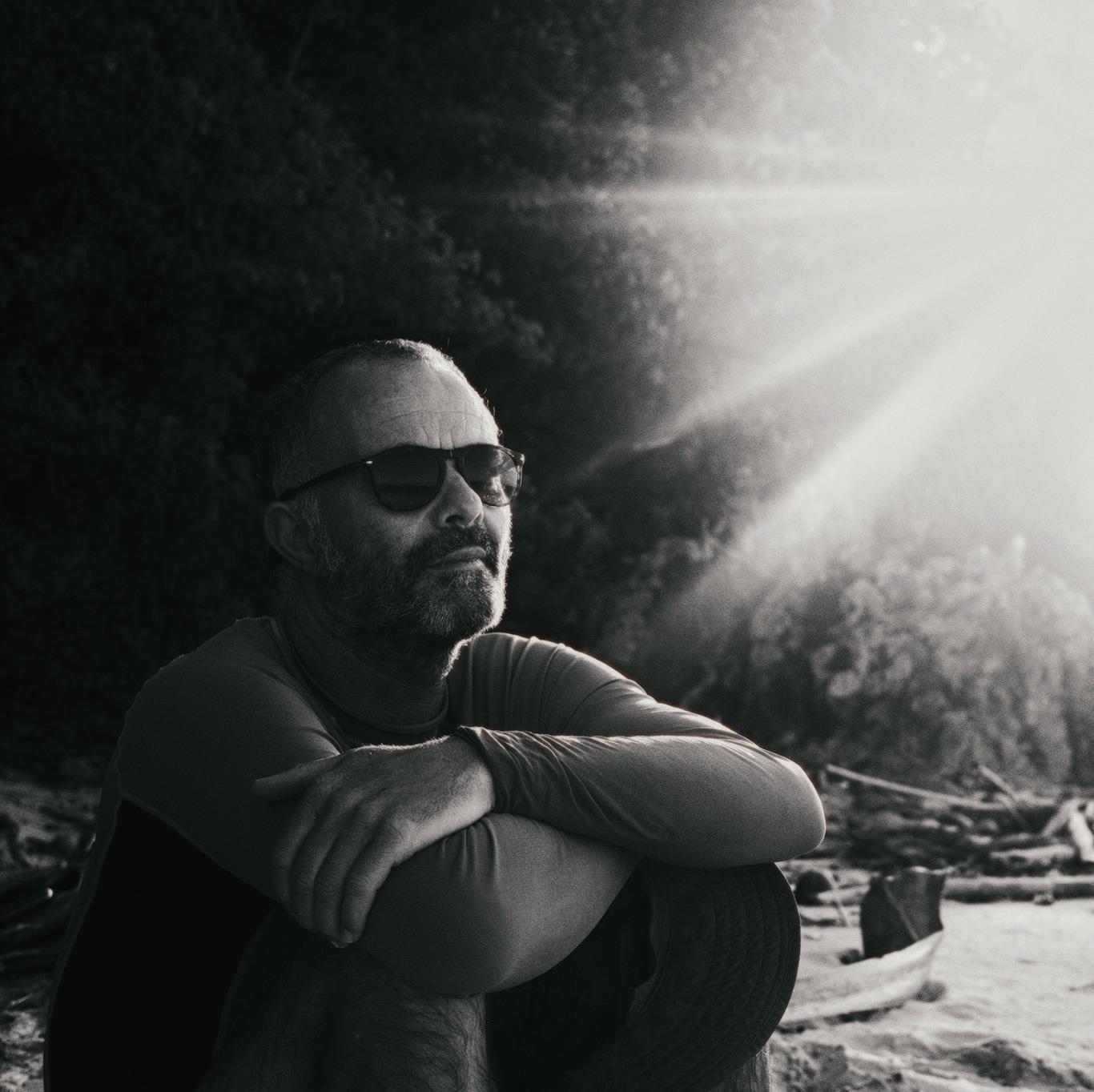 Black and white portrait of Nharyan, arms crossed, backlit on a beach