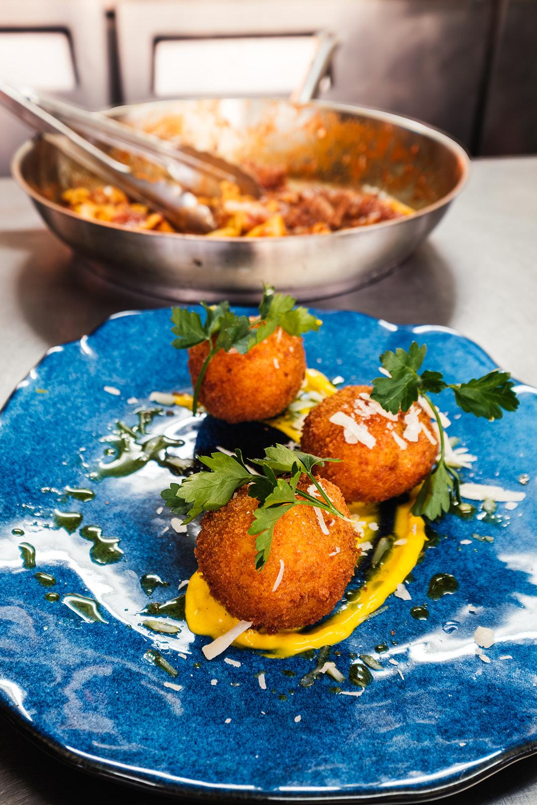 Three arancini balls on a hand-glazed blue plate, a pan of ragu in the background