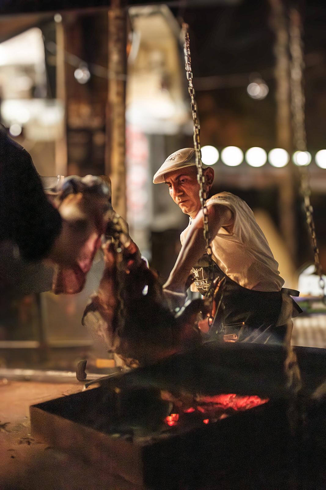 A chef tending meat roasting on chains over an open fire pit at night