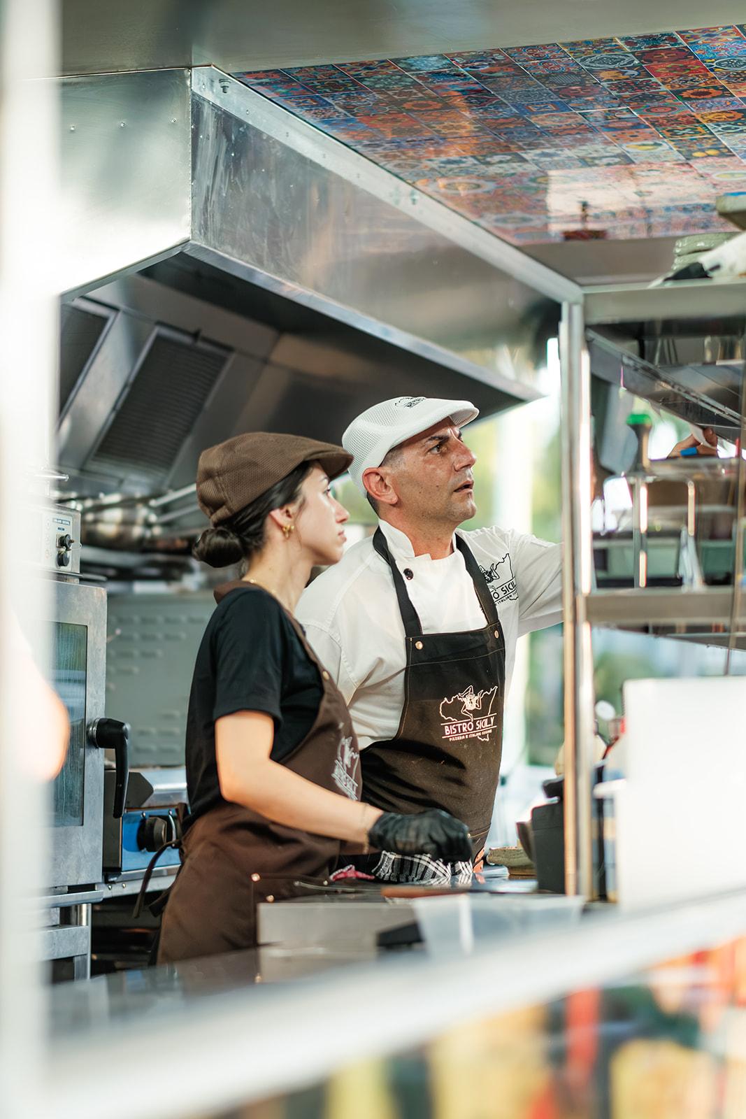 Two Bistro Sicily staff in branded aprons working the truck's service window, colourful tiled ceiling above