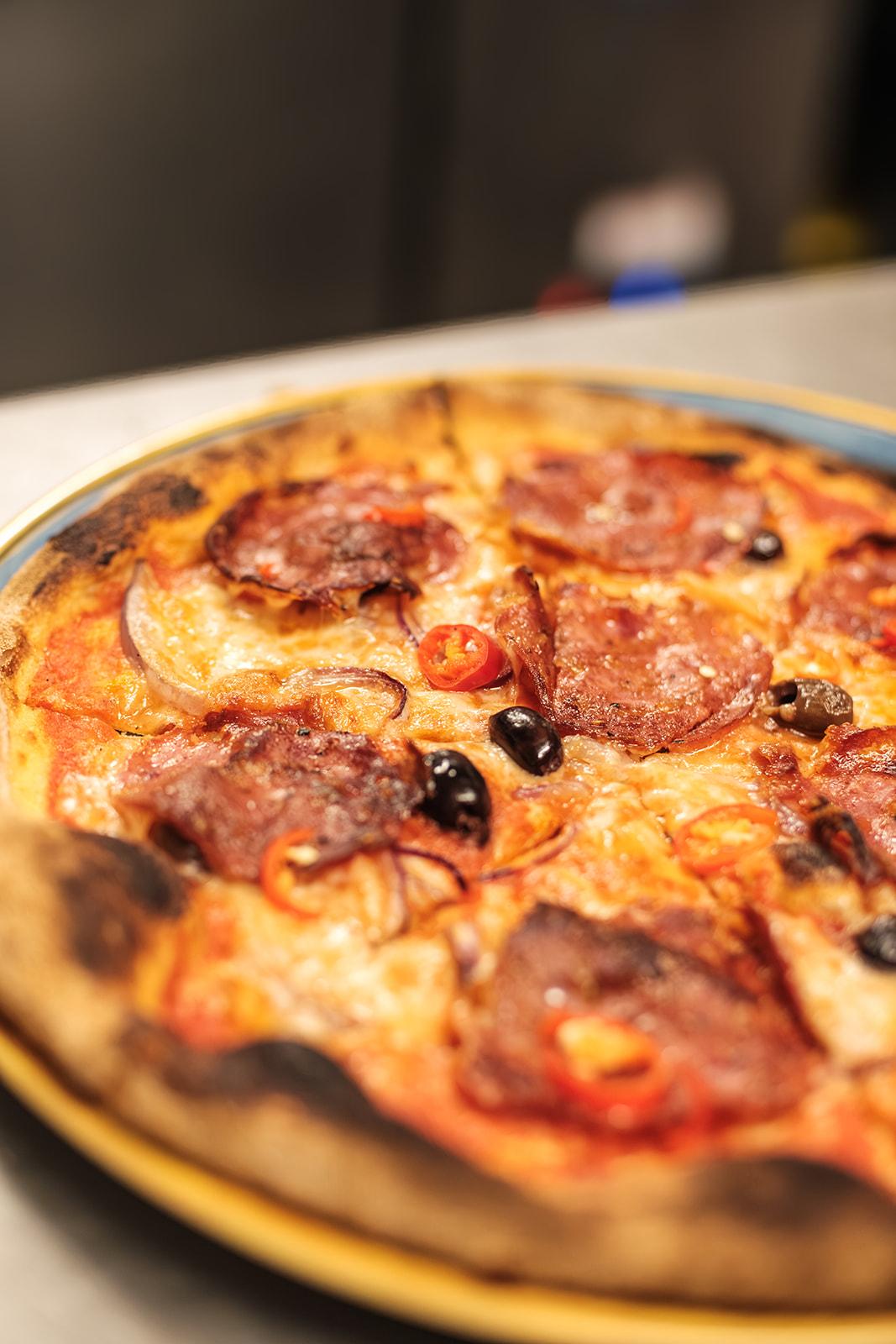 Close-up of a wood-fired pizza topped with salami, chilli and olives