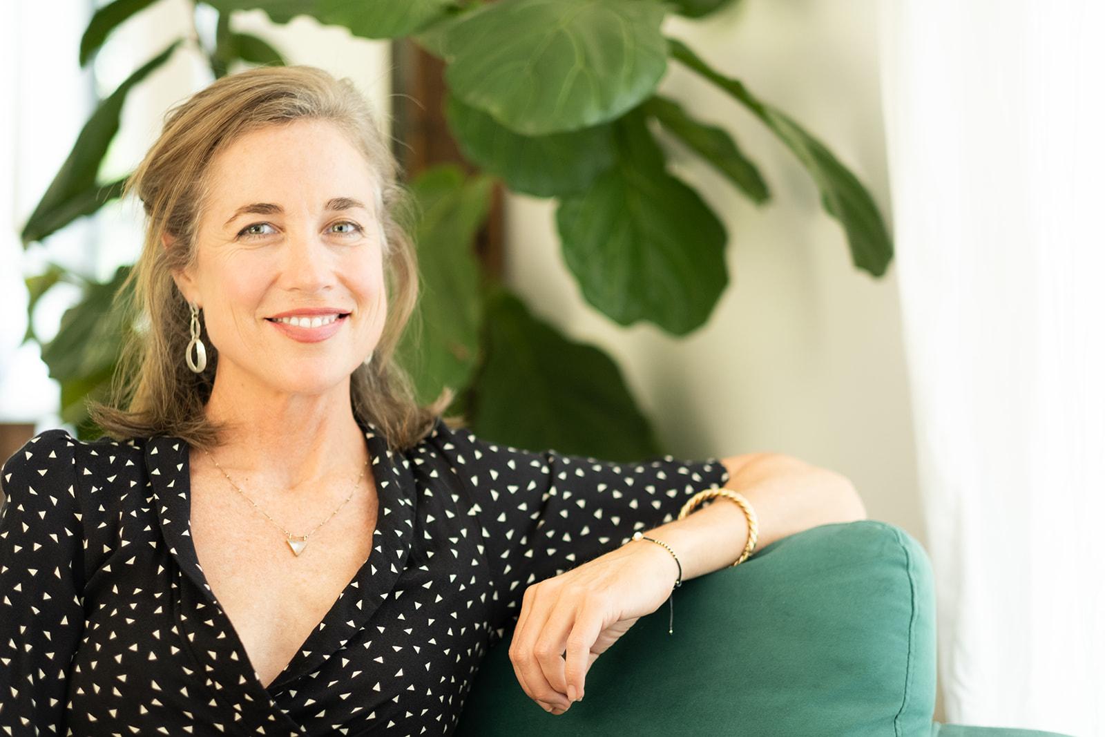 Grace Hawkins seated beside a fiddle-leaf fig, resting an arm on a green velvet chair