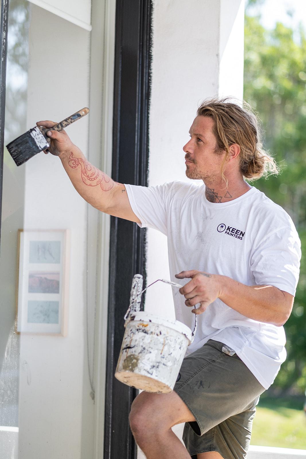 A painter reaching up with a brush and paint tin, one knee braced on a ladder step