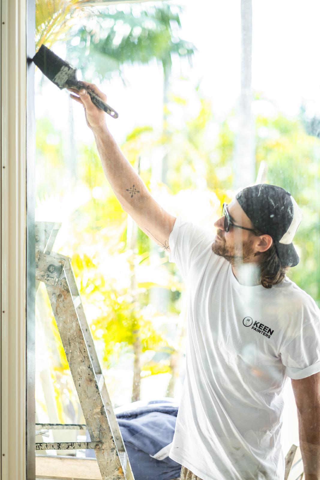 A painter reaching up to a high window frame, tropical greenery blurred behind