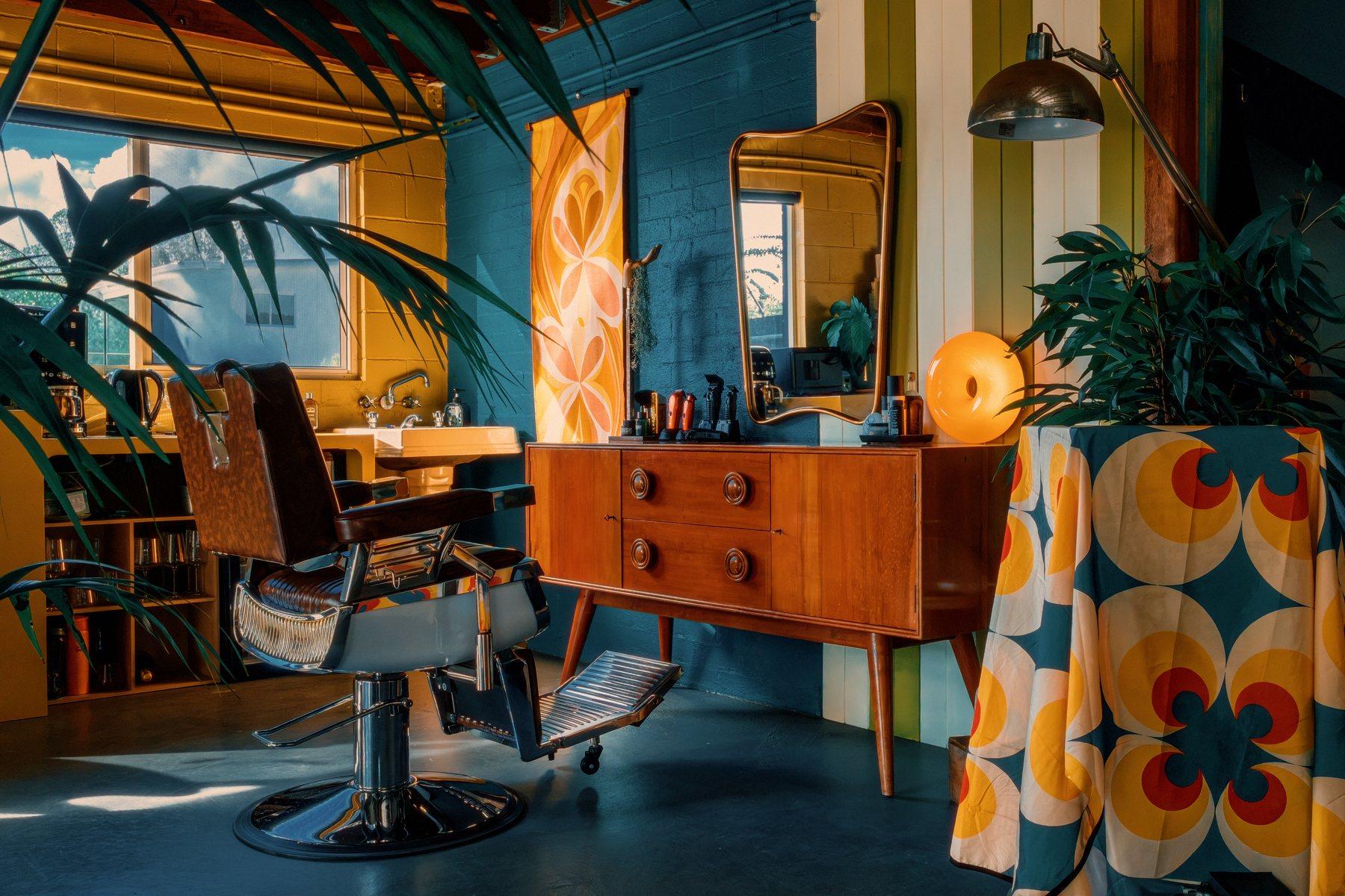 Barber chair beside a mirror and dresser, orange lamp glowing, plants either side