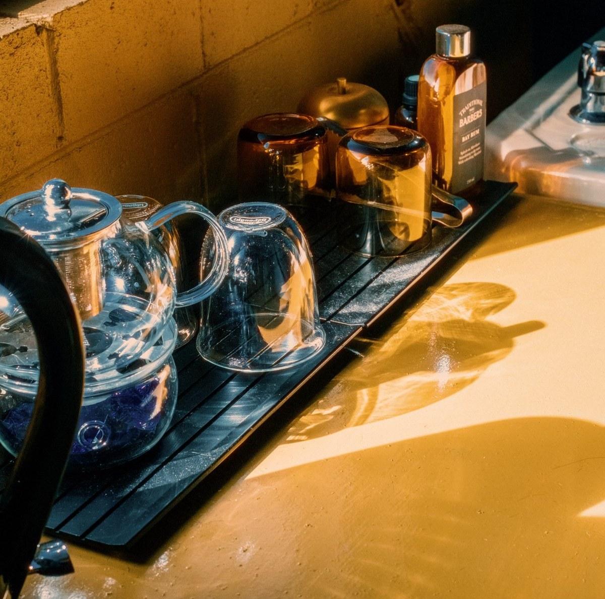 A glass teapot and cups beside a bottle of bay rum on the wash basin's edge, catching afternoon light