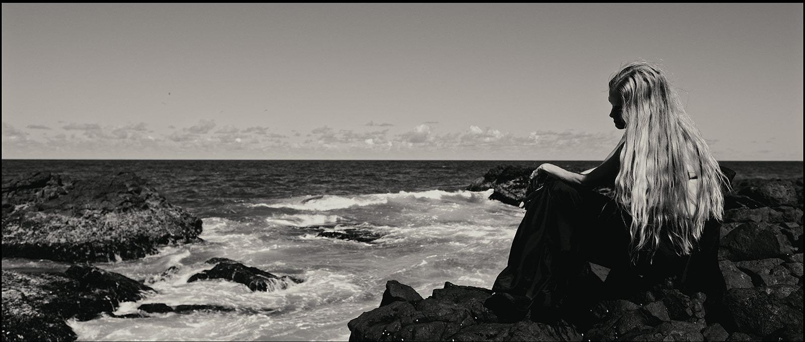 A woman crouched on the rocks, hair blowing in the wind, black and white