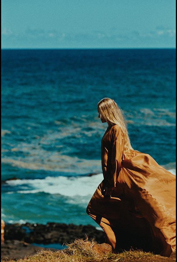 A woman in the flowing copper dress walking toward the turquoise water's edge