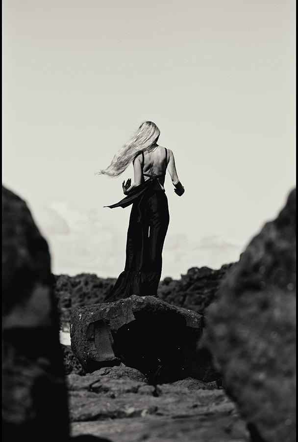 A woman seen from behind, hair blowing sideways between rock formations, black and white
