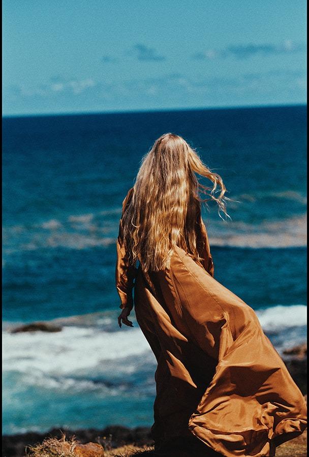 A woman in the flowing copper dress looking back over her shoulder toward the ocean
