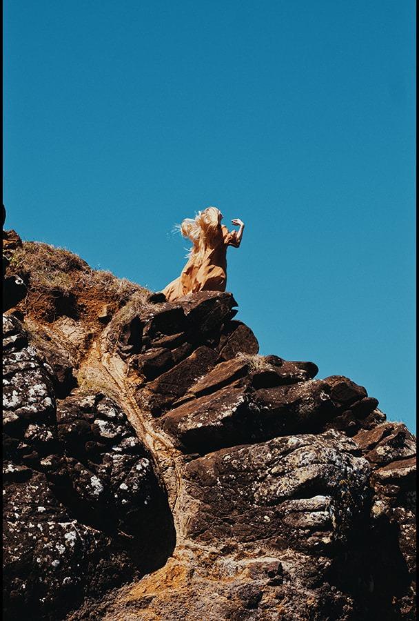 A woman in the flowing copper dress perched on a rock spire, arm raised against the sky