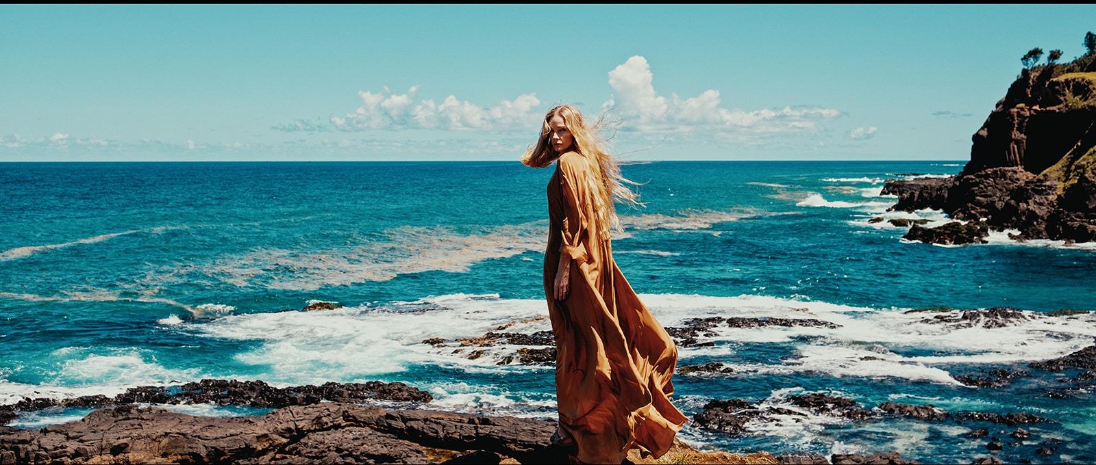 A woman in a flowing copper dress standing on a rocky point above the ocean