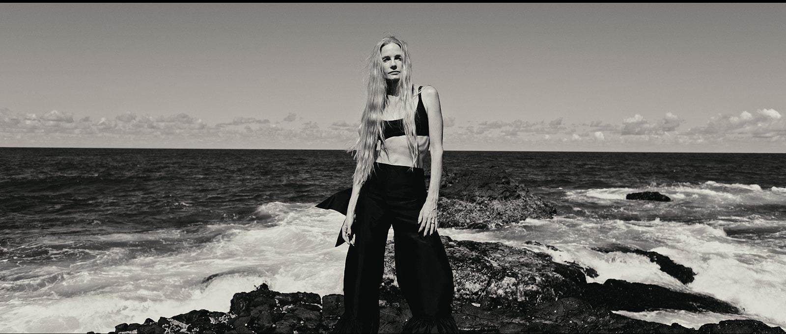 A woman in black standing on the rocks as waves break behind her, black and white