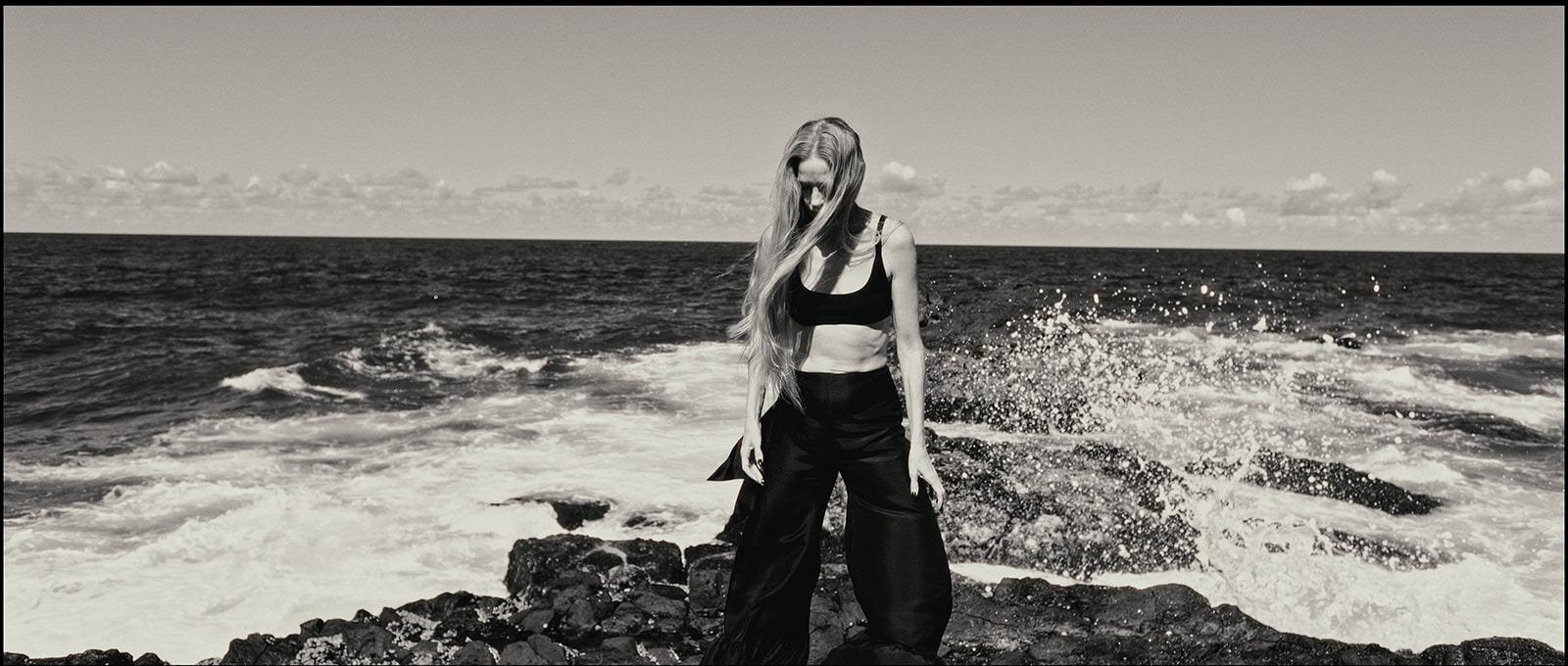 A woman in black standing on the rocks as a wave breaks beside her