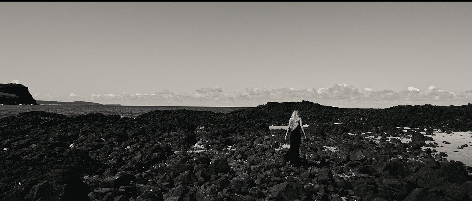 A wide, moody view of the rocky coastline, a distant figure barely visible, black and white