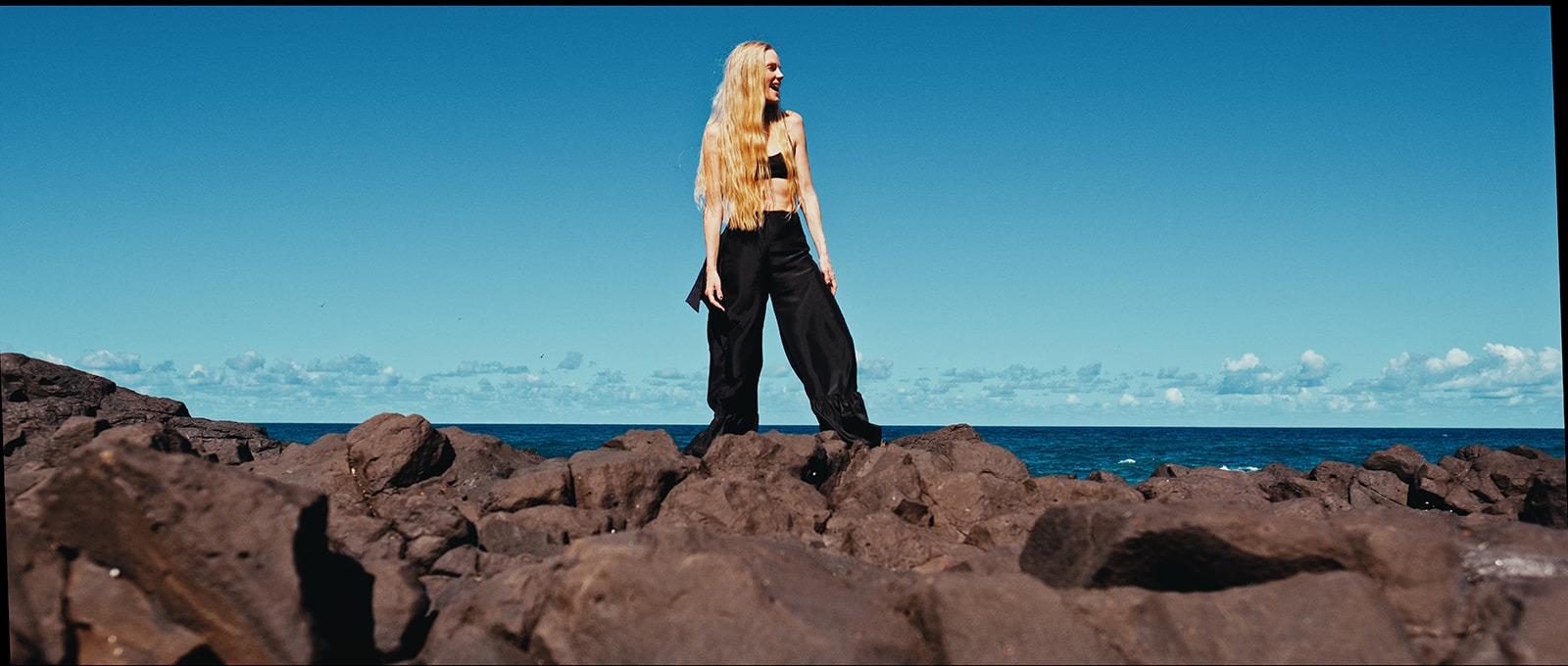 A woman in black standing on the rocks against a wide blue sky