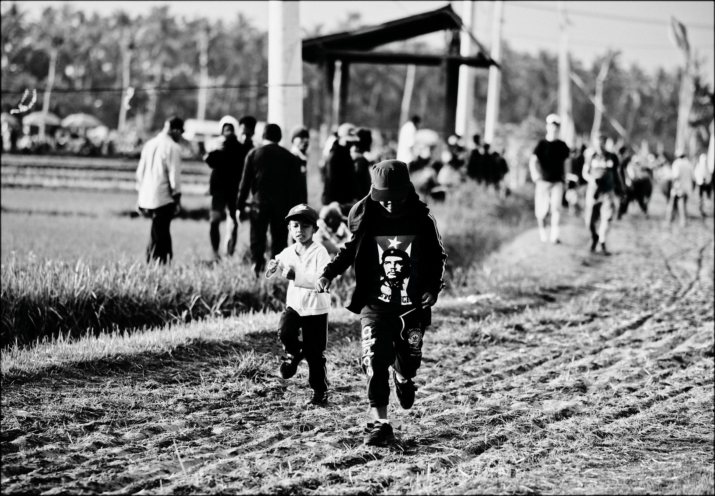 Black and white: a crowd walking along a path through a field toward a stage, a child running in the foreground