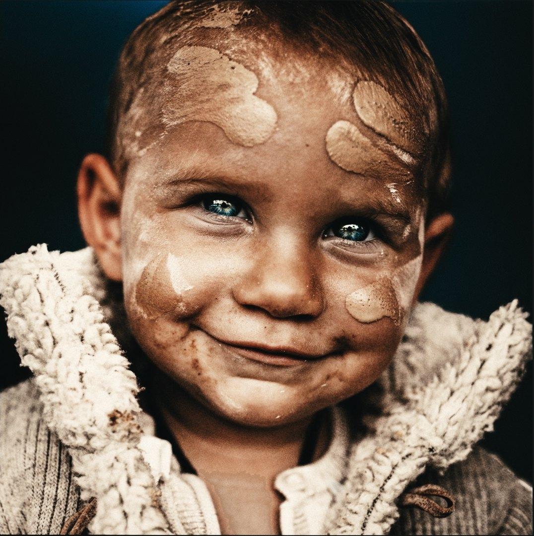 A smiling toddler with blue eyes and an ochre-painted face, in a cream woolly jacket