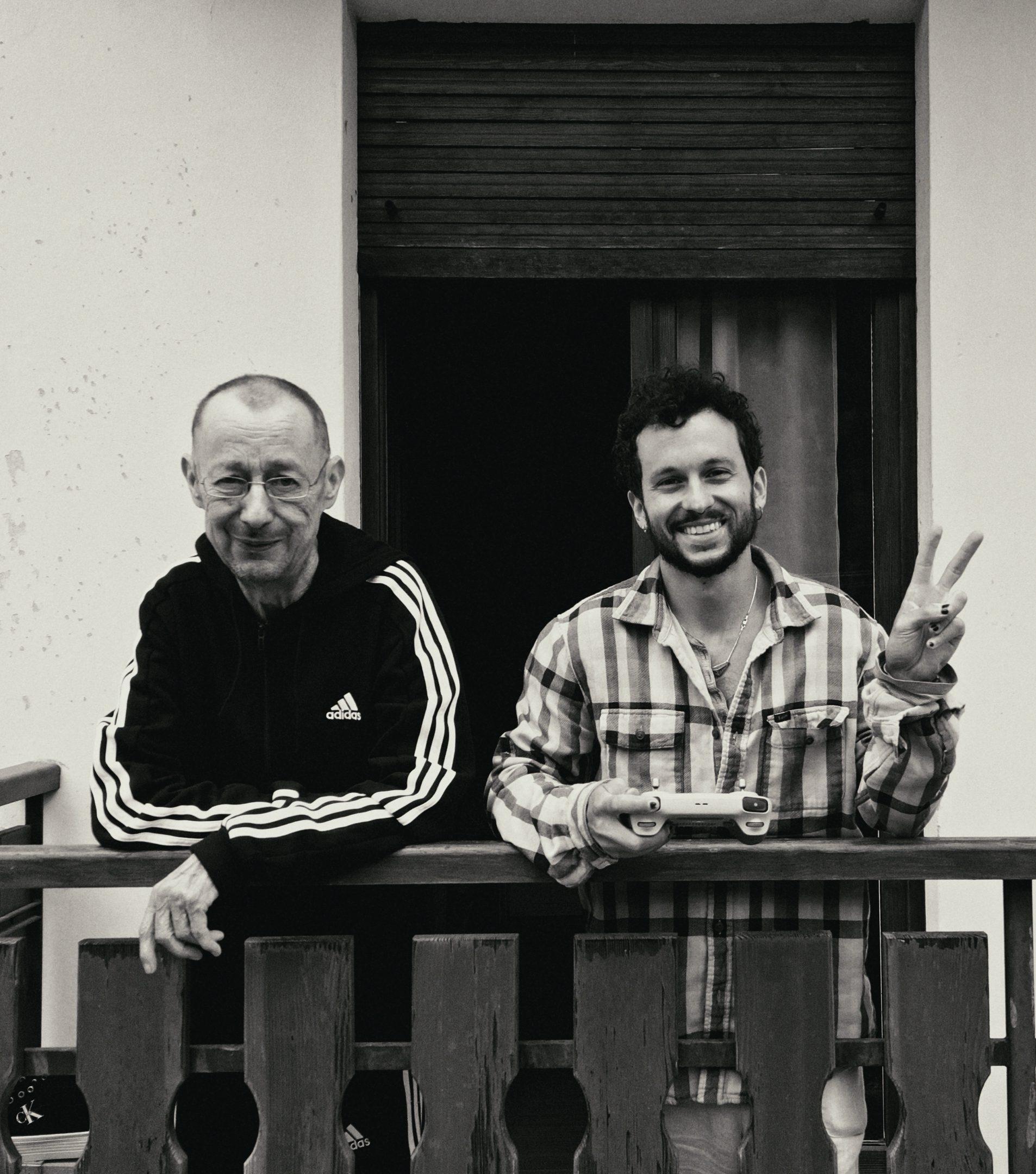 Black and white: two men leaning on a wooden balcony rail, one giving a peace sign to the camera