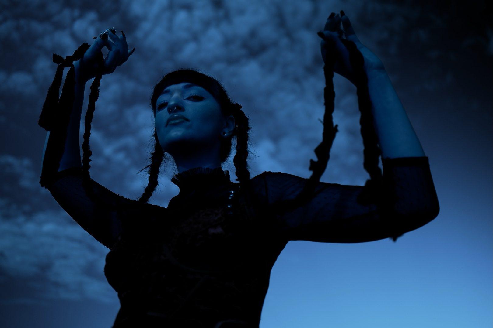 Juman against a deep blue sky, arms raised and holding her braids