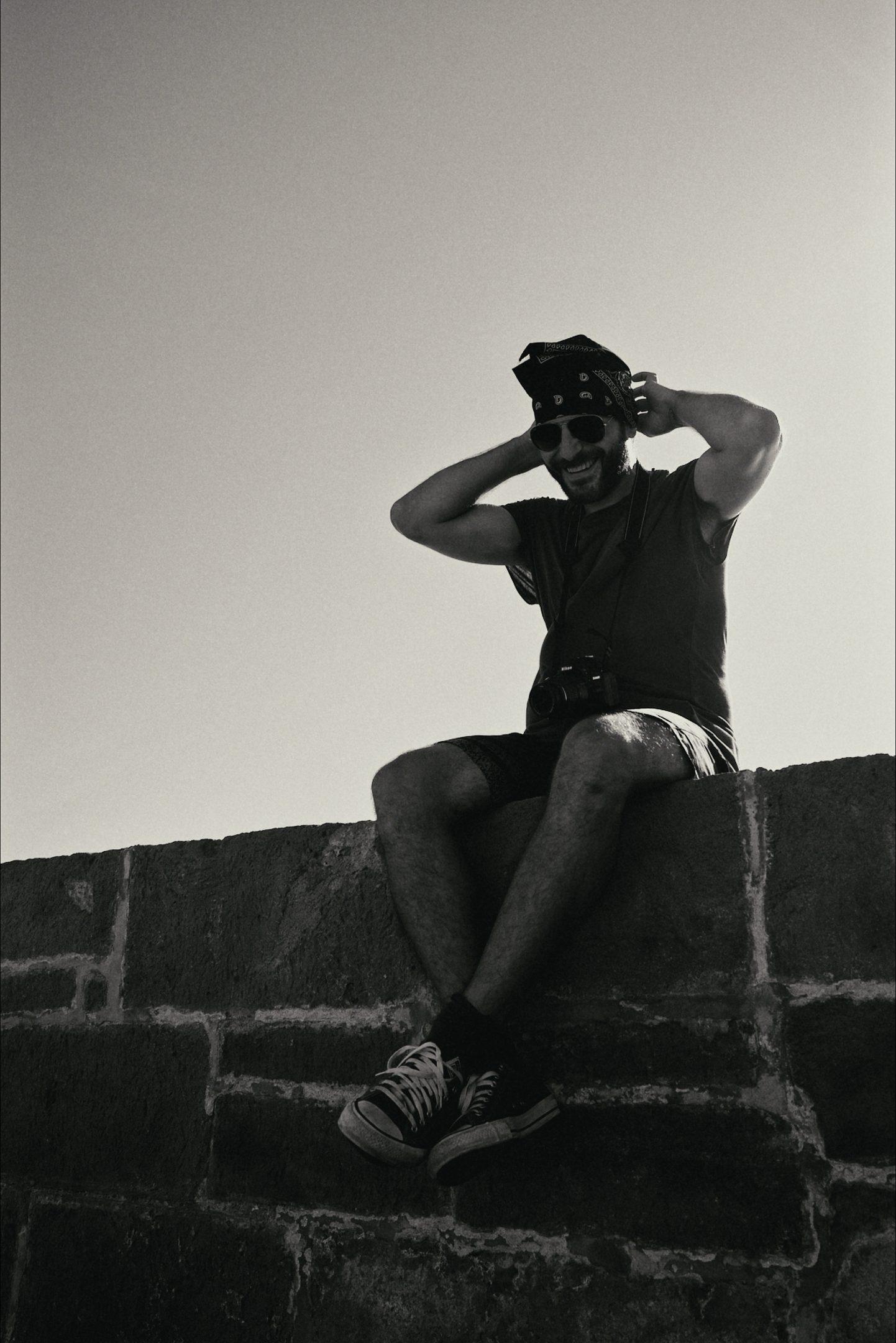 A man sitting on an old stone wall with his hands on his head, against a pale sky