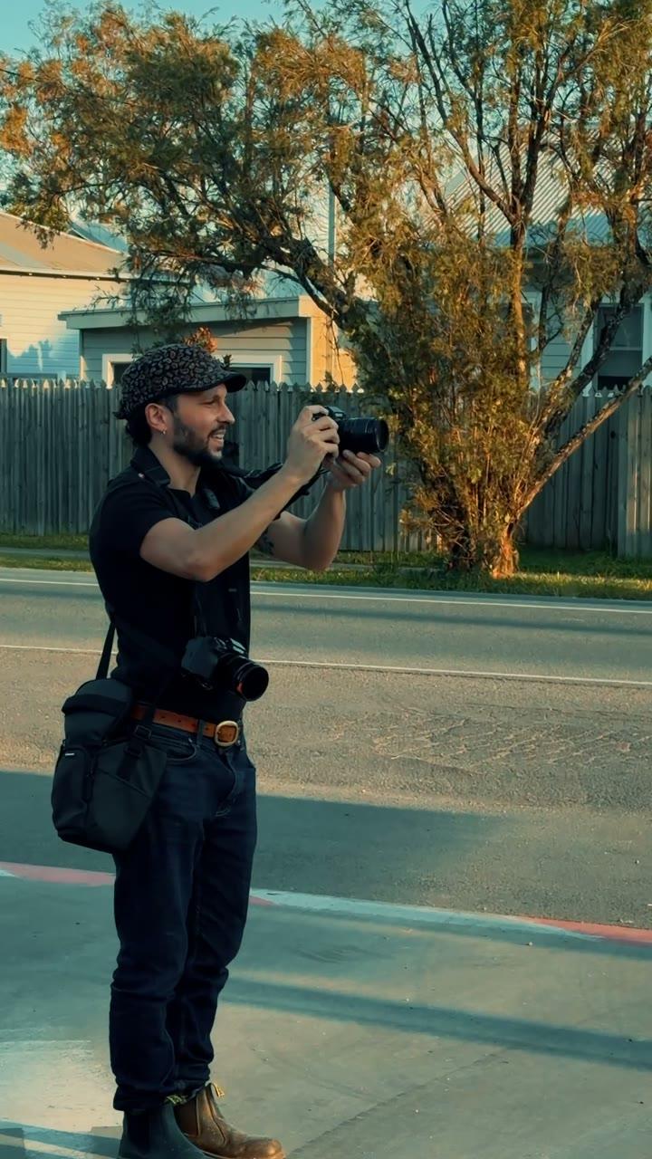 A man in a patterned cap photographing the street with a camera, a second camera on his chest, in low golden light