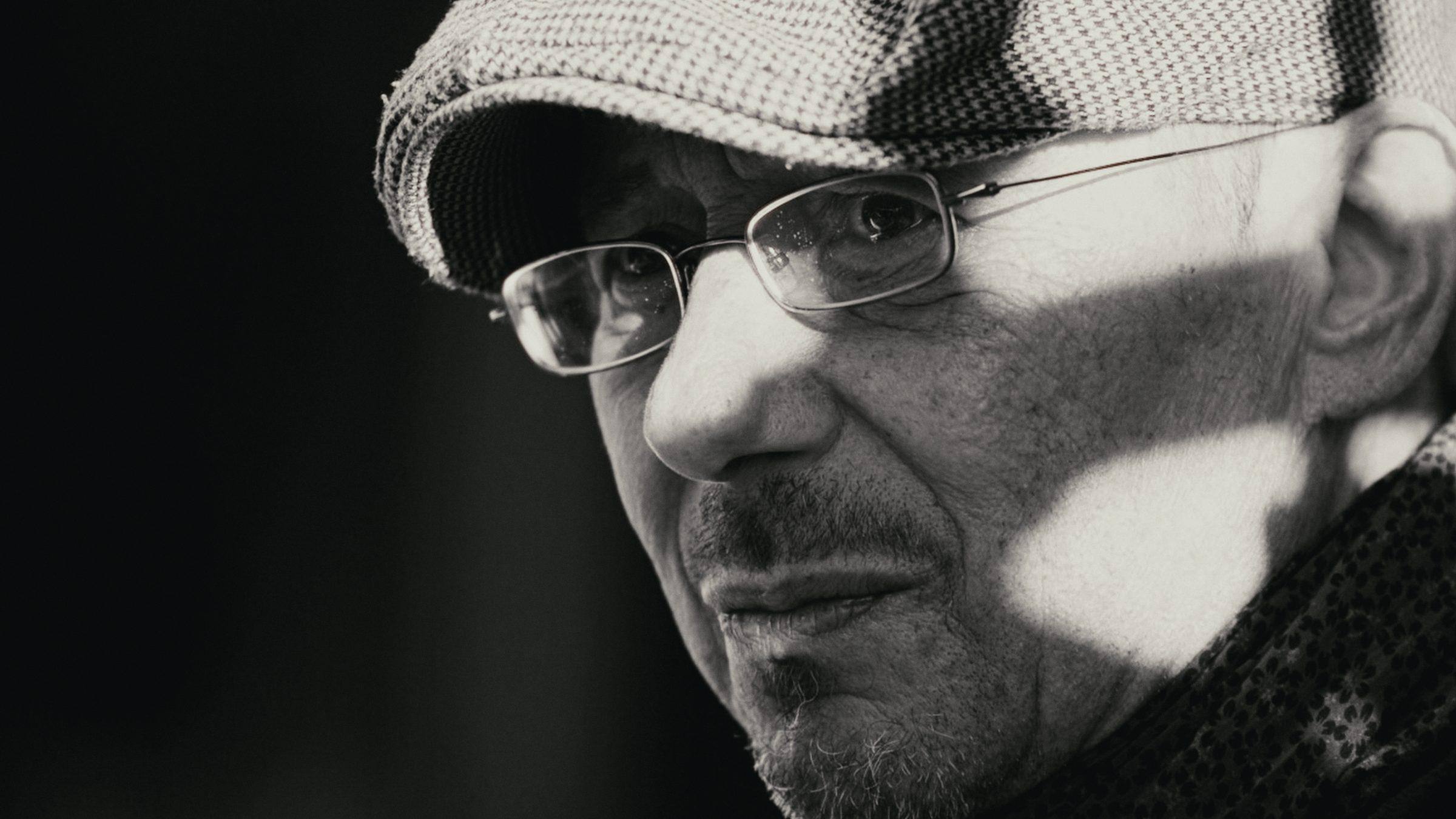 Black and white portrait of an older man in a flat cap and glasses, a shadow of sunlight across his face