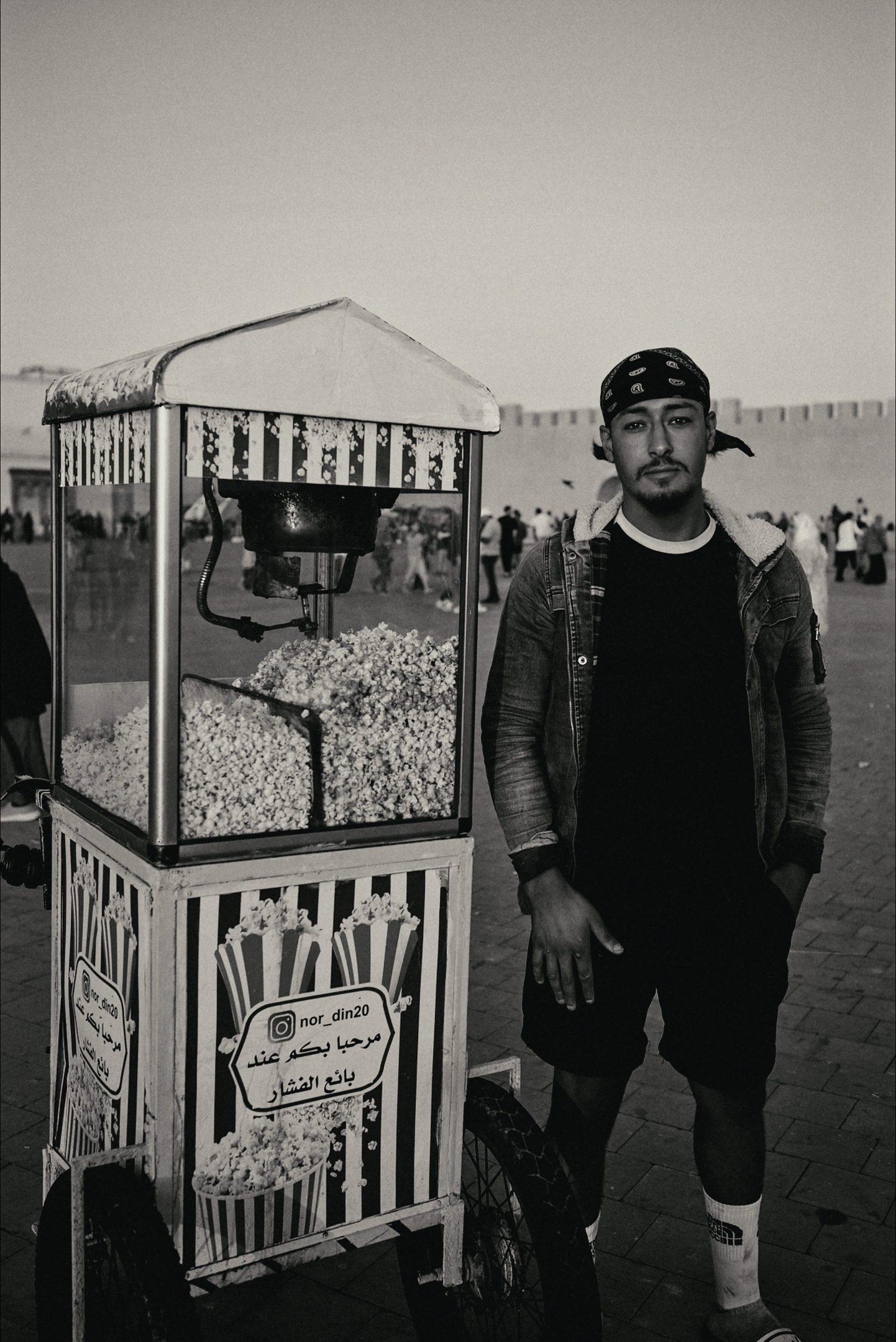 Black and white: a man standing beside a vintage popcorn cart in a town square