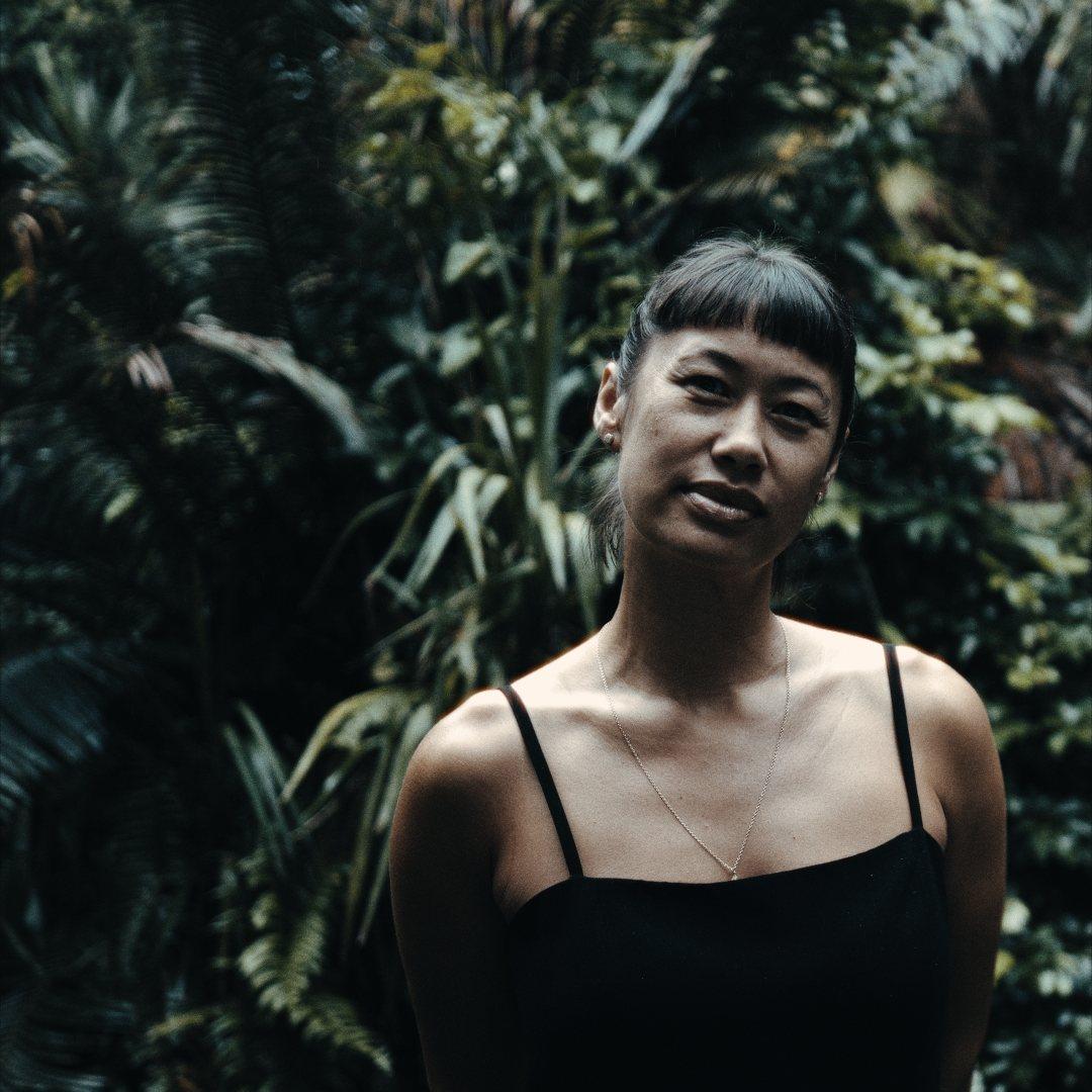 A woman with a fringe in a black strappy top, standing in front of dark green foliage