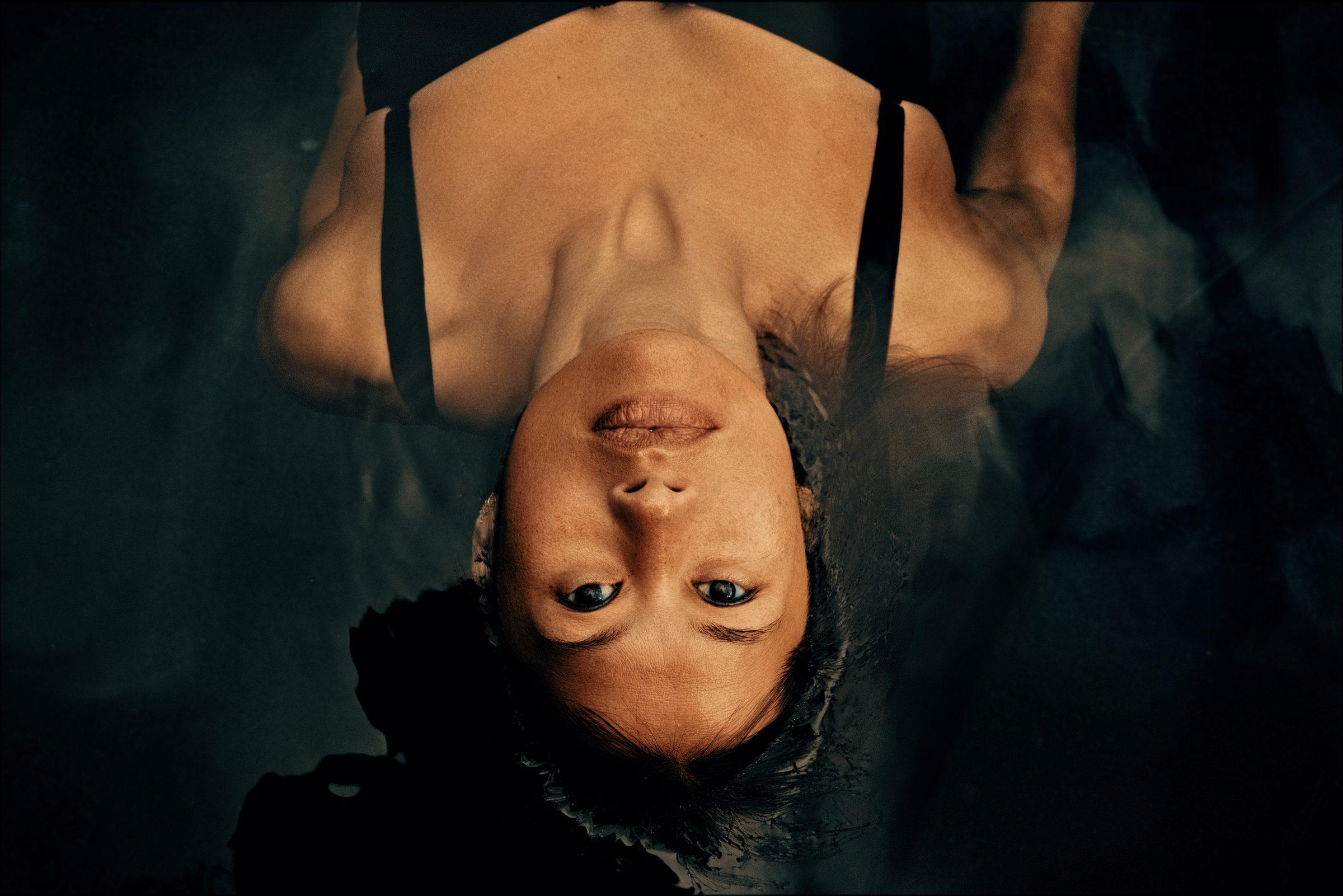 An upside-down portrait of a woman in dark clothing, lit in blue