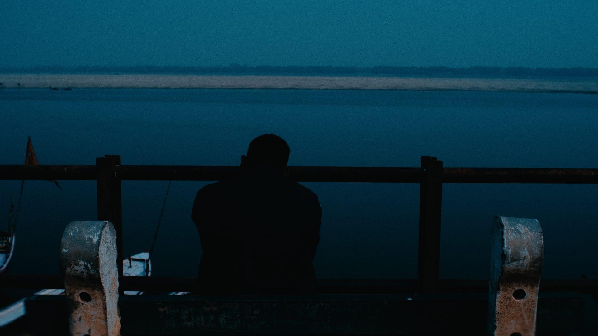 A figure silhouetted at a railing over dark blue water at dusk