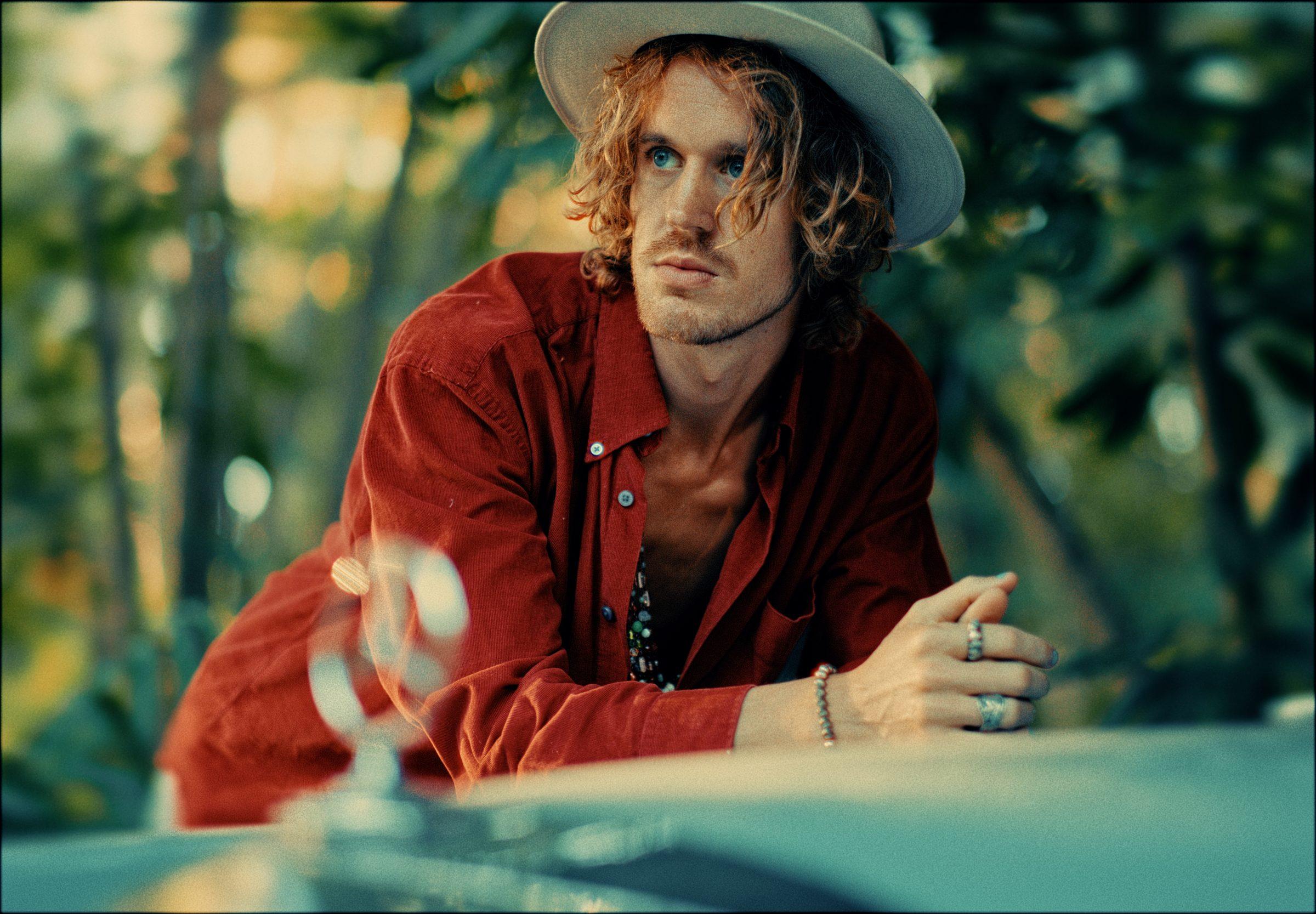 Kenzo Cruz in a cream hat and red shirt, leaning on the bonnet of a car among trees