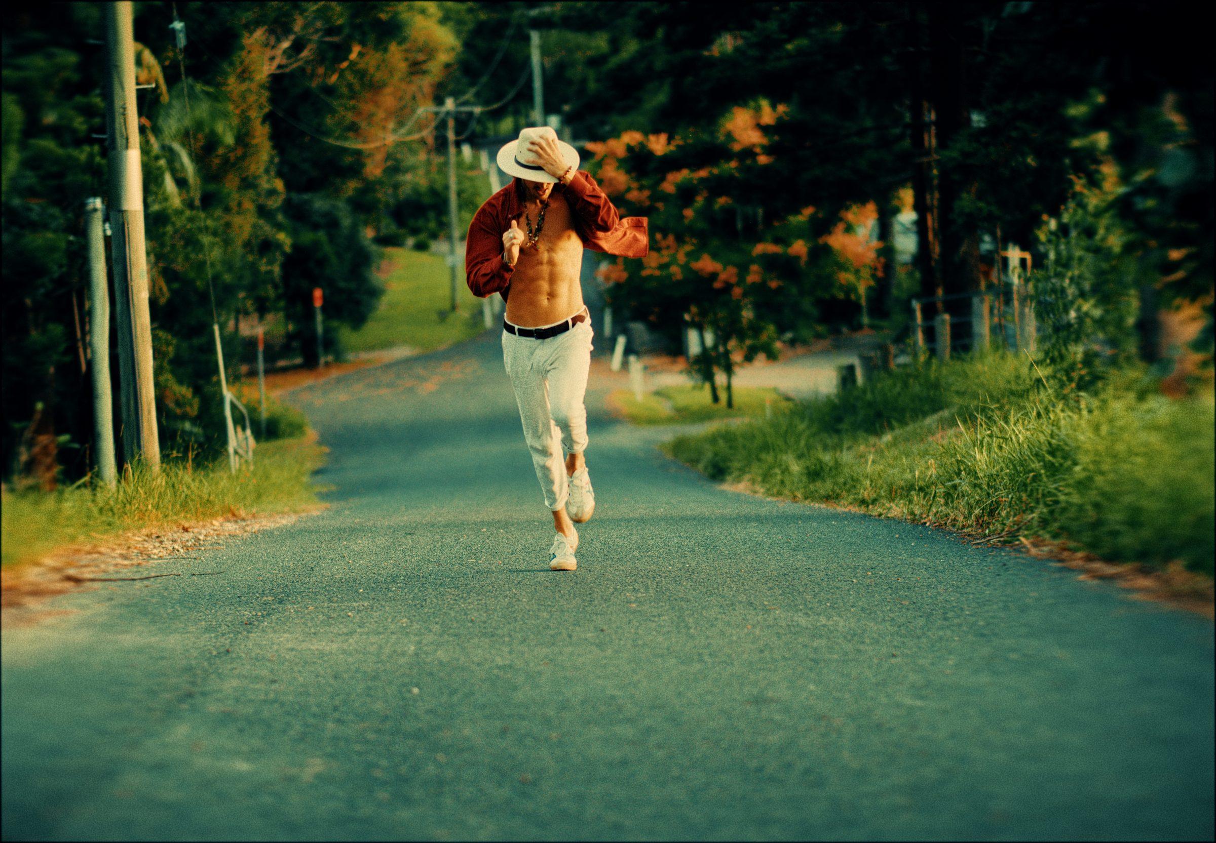 Kenzo Cruz walking down a tree-lined road in a cream hat, shirt open