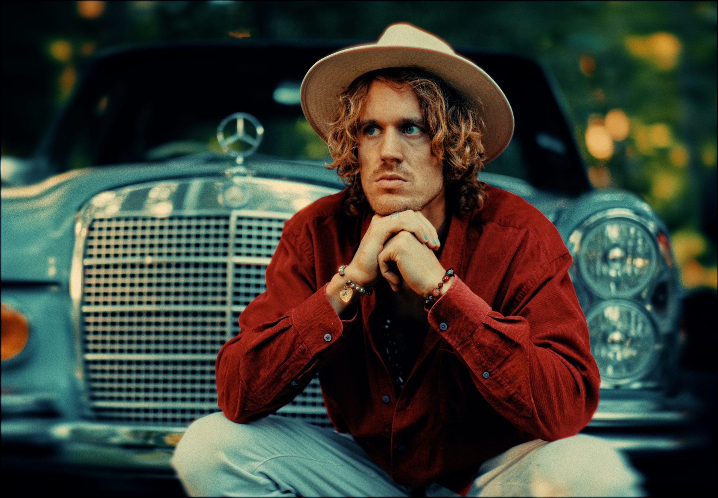 Kenzo Cruz crouched in front of a vintage Mercedes, hands clasped under his chin, in a cream hat and red shirt