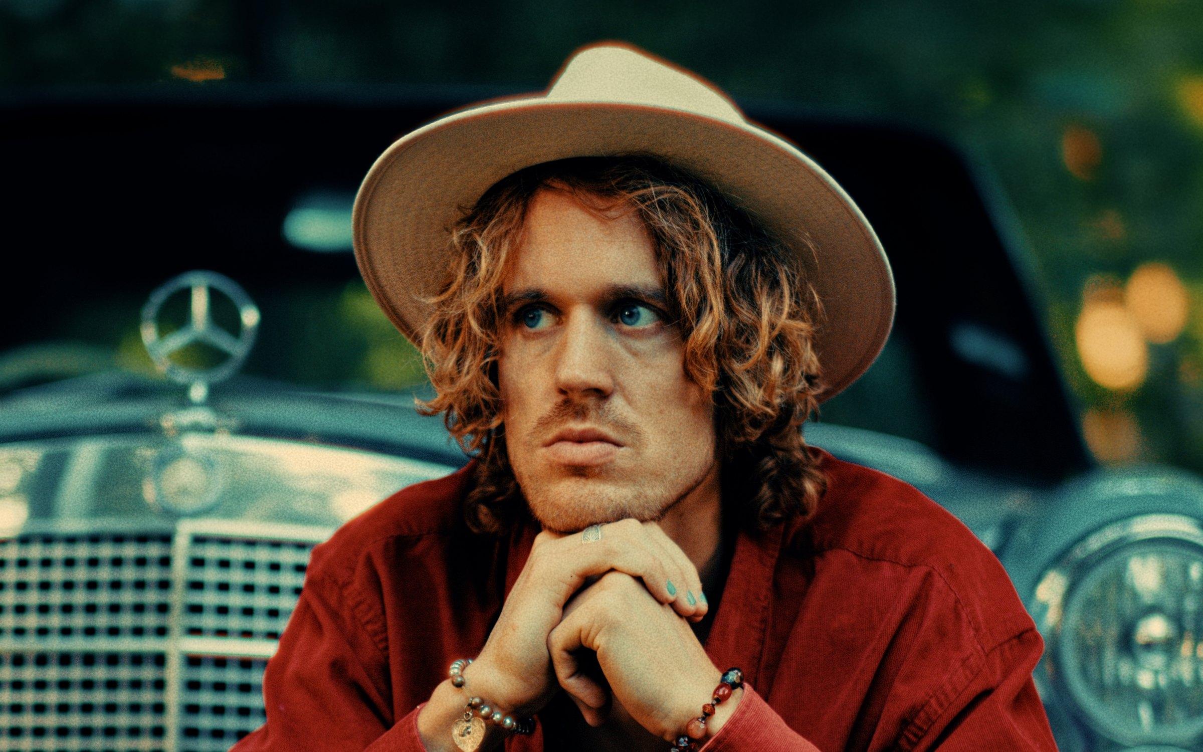 Kenzo Cruz, close on his face under a cream hat, hands clasped under his chin, a vintage Mercedes behind him