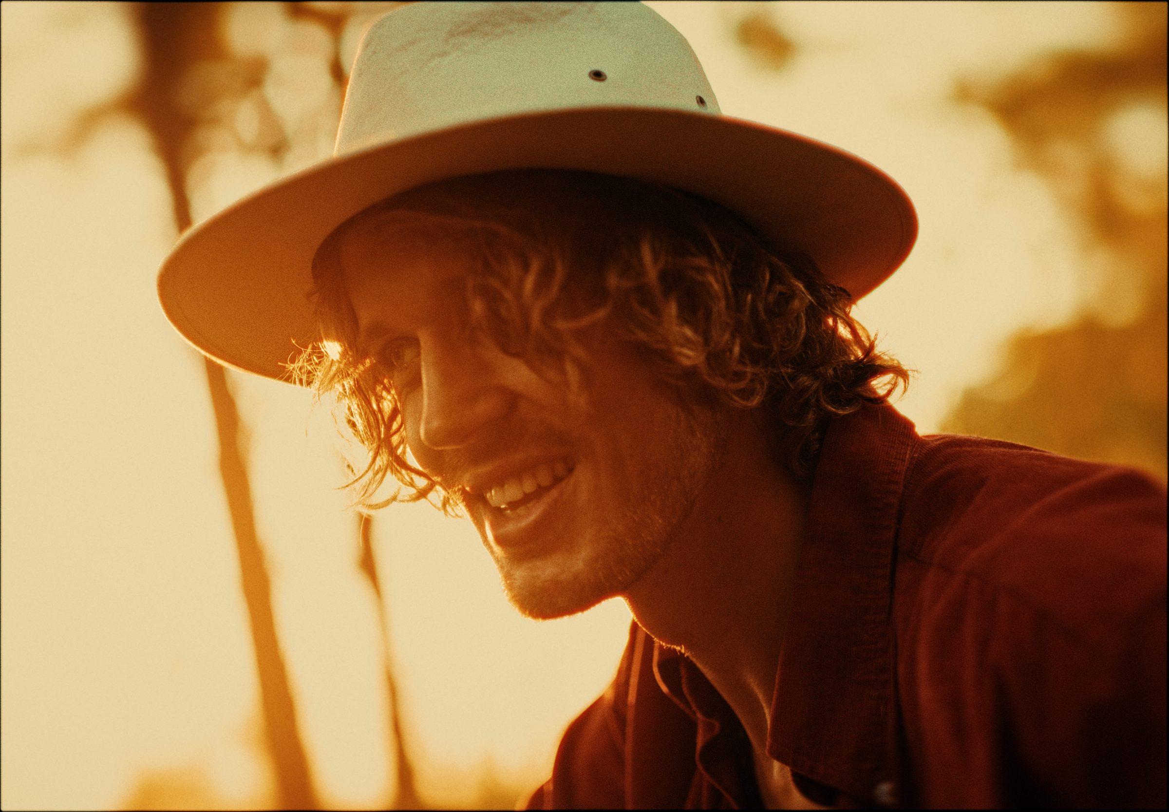 A close portrait of Kenzo Cruz laughing in golden light, in a cream hat
