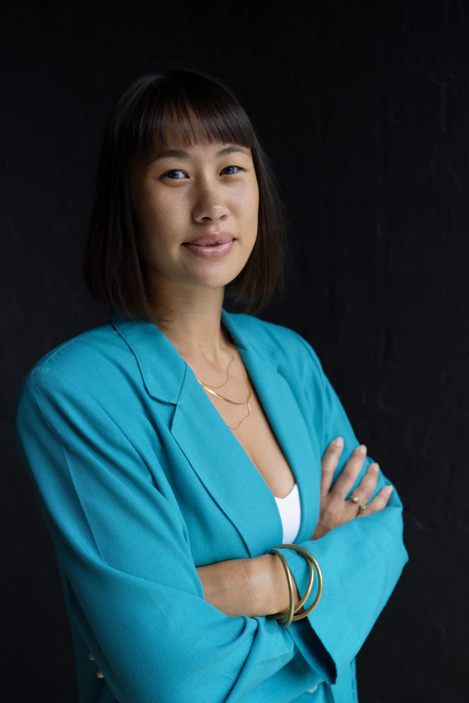 Roxy, headshot against a dark background, in a bright teal blazer with her arms crossed