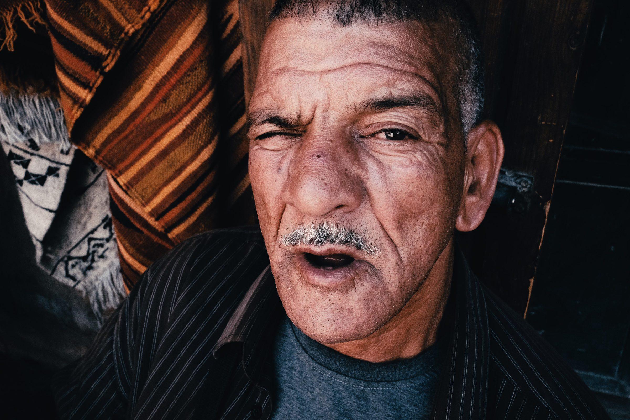 Close portrait of a man with a grey moustache, one eye narrowed, in a dim shop hung with woven textiles