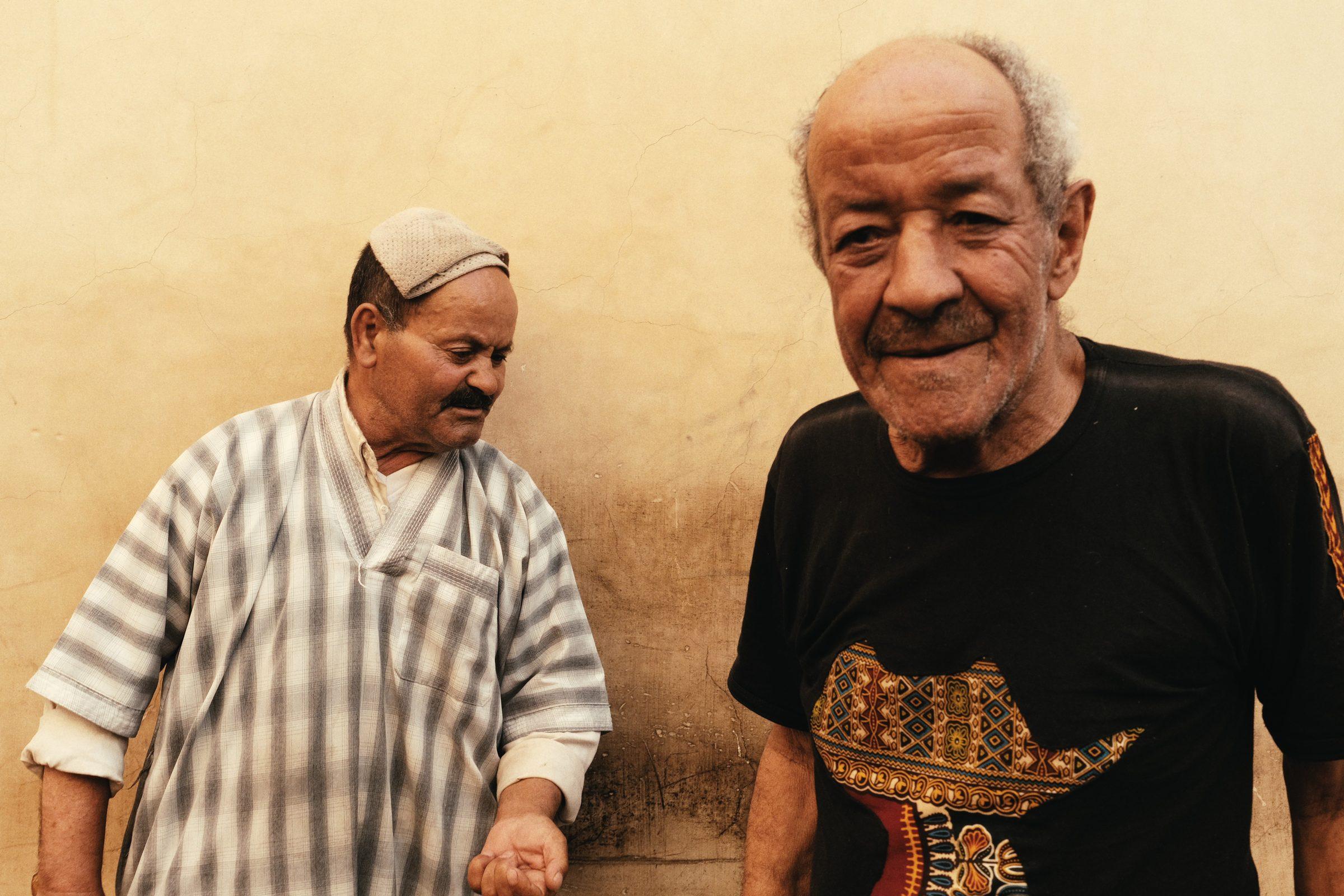 Two older men side by side against an ochre wall, one in a striped robe and cap, the other smiling in a black T-shirt