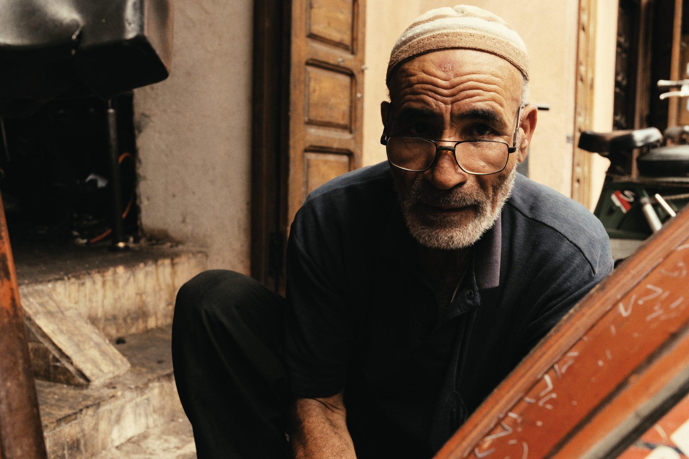 An older man with glasses and a white beard, in a skullcap, crouched in a doorway beside an orange-painted cart