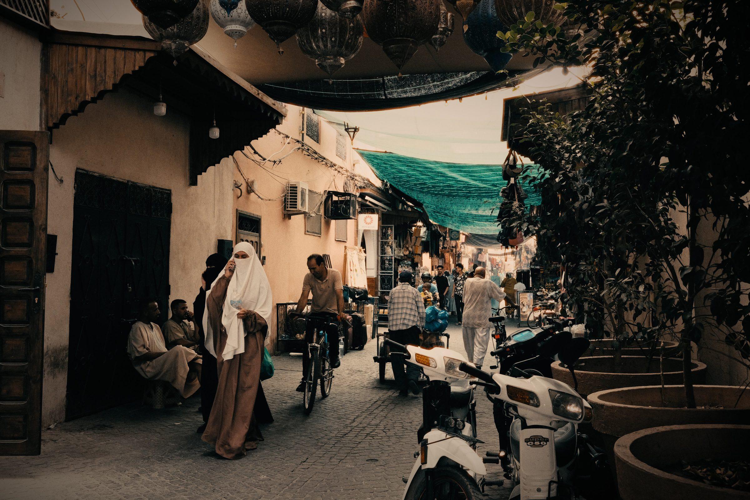 A narrow Marrakesh alley: a woman in white walking past scooters and a cyclist, lanterns and a green awning overhead