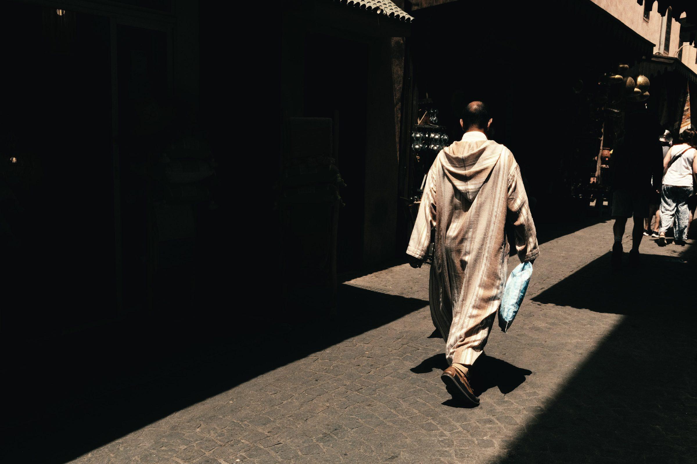 A man in a long pale robe walks through a stripe of sunlight in a dark, covered street