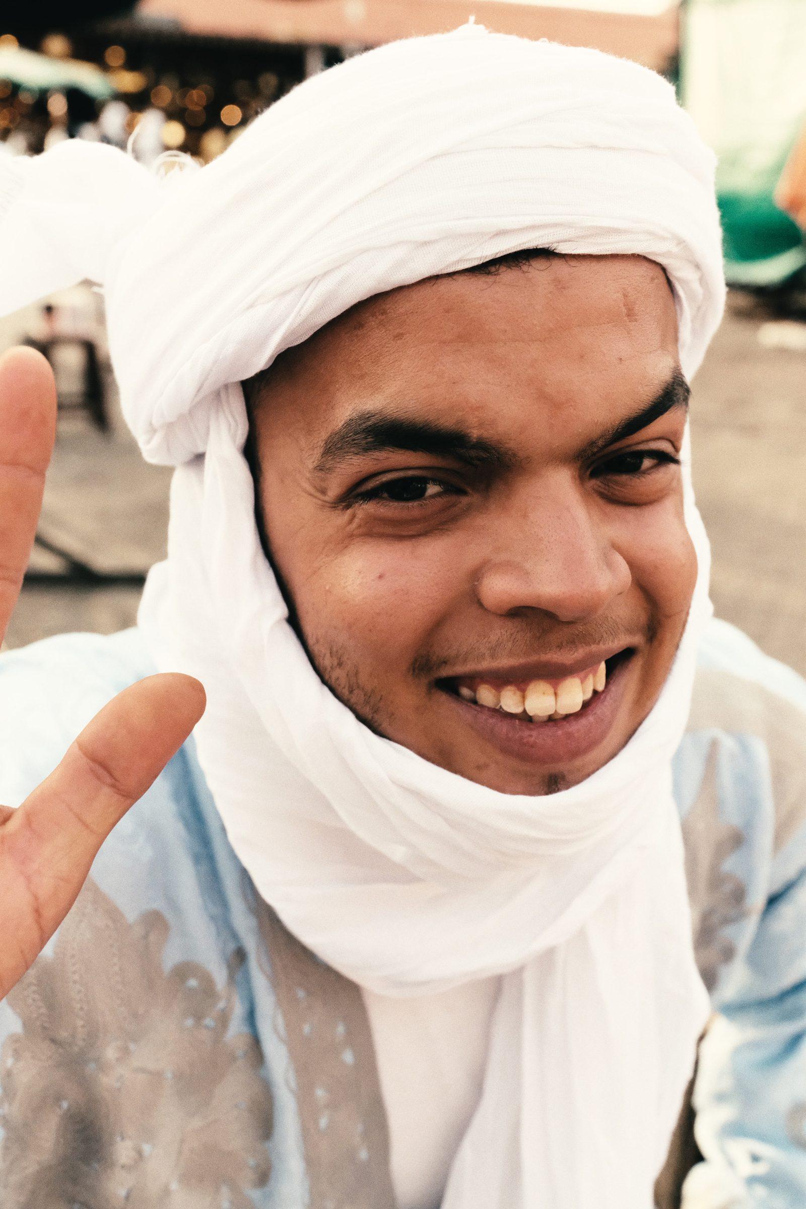 A smiling young man wrapped in a white turban and scarf, thumb raised