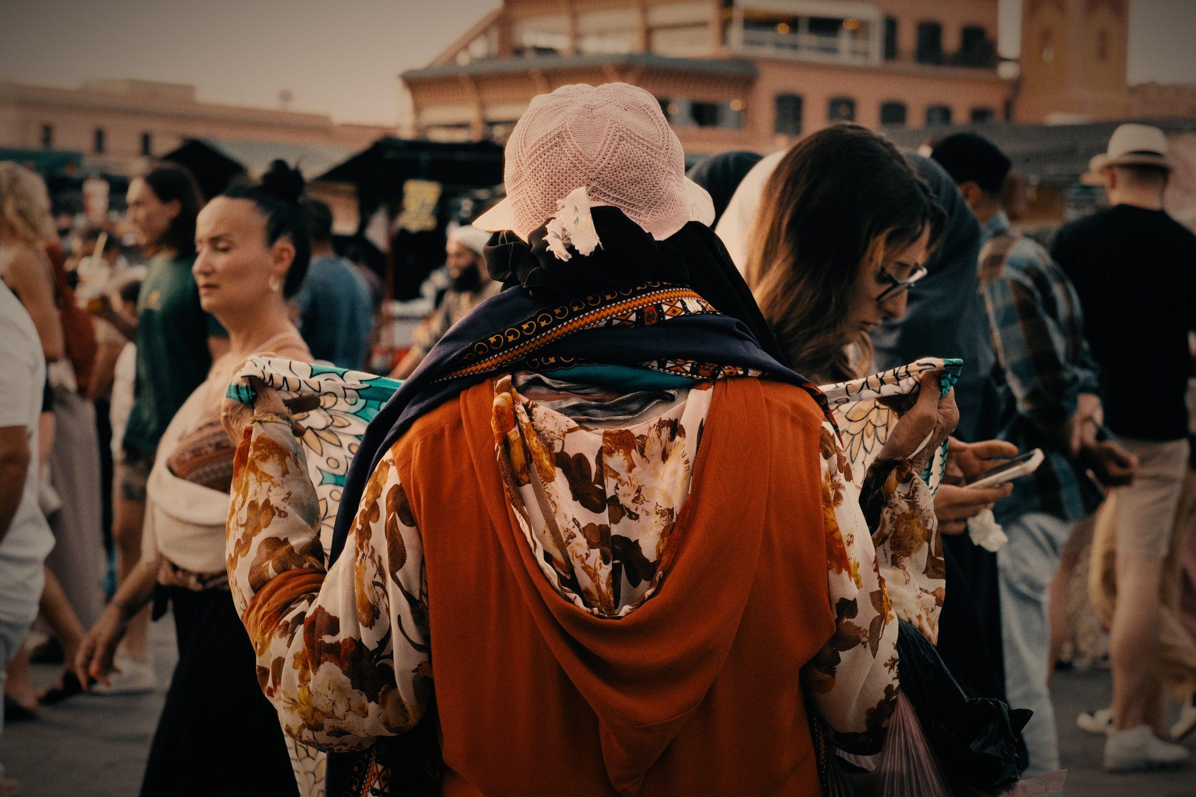 A crowd in the square, a woman in an orange and floral hooded robe turned away from the camera