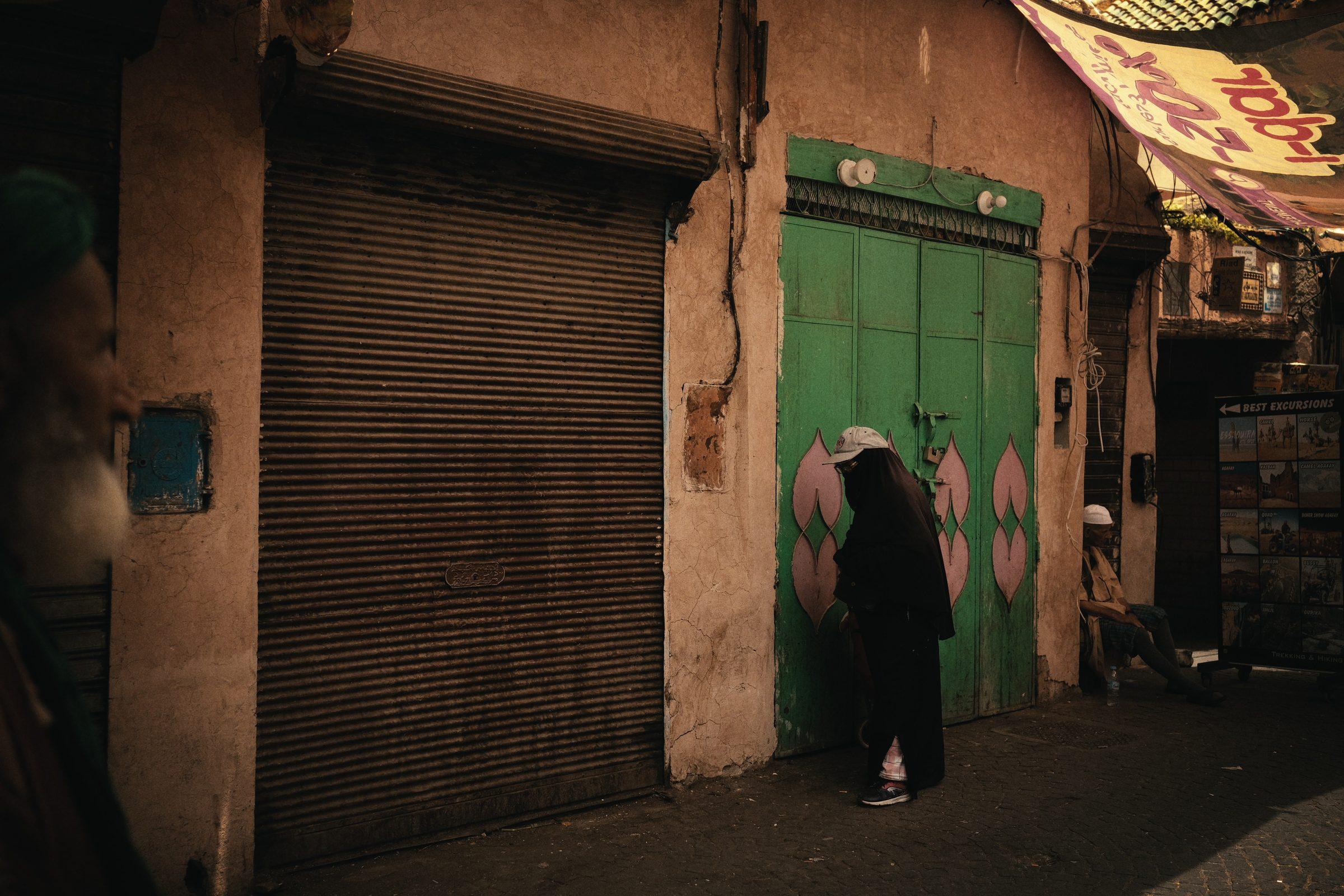 A man bent in front of a green door painted with pink flowers, beside a closed roller shutter