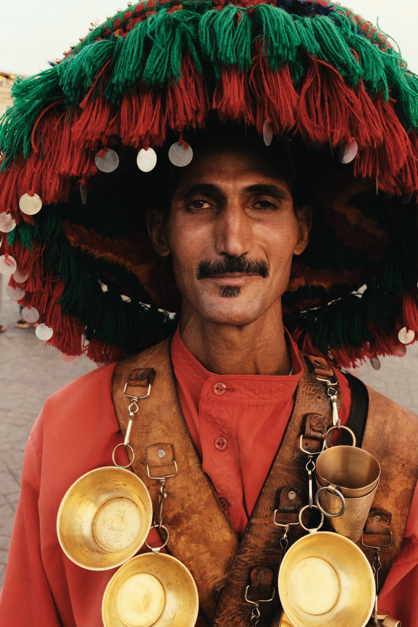 A water seller in a tasselled red and green hat, brass cups across his chest, looking at the camera