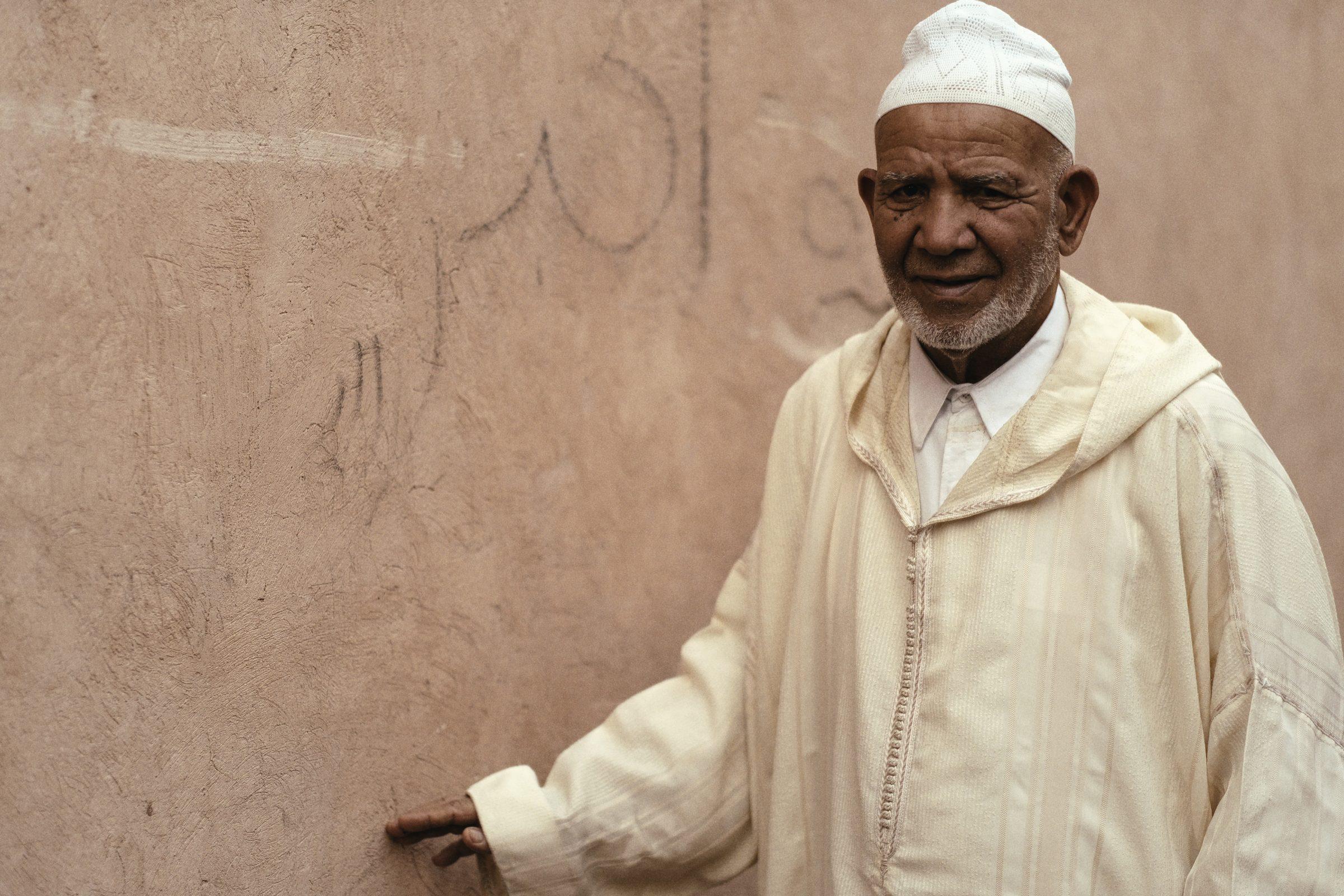 An elderly man in a white robe and cap resting a hand on a weathered wall, looking at the camera
